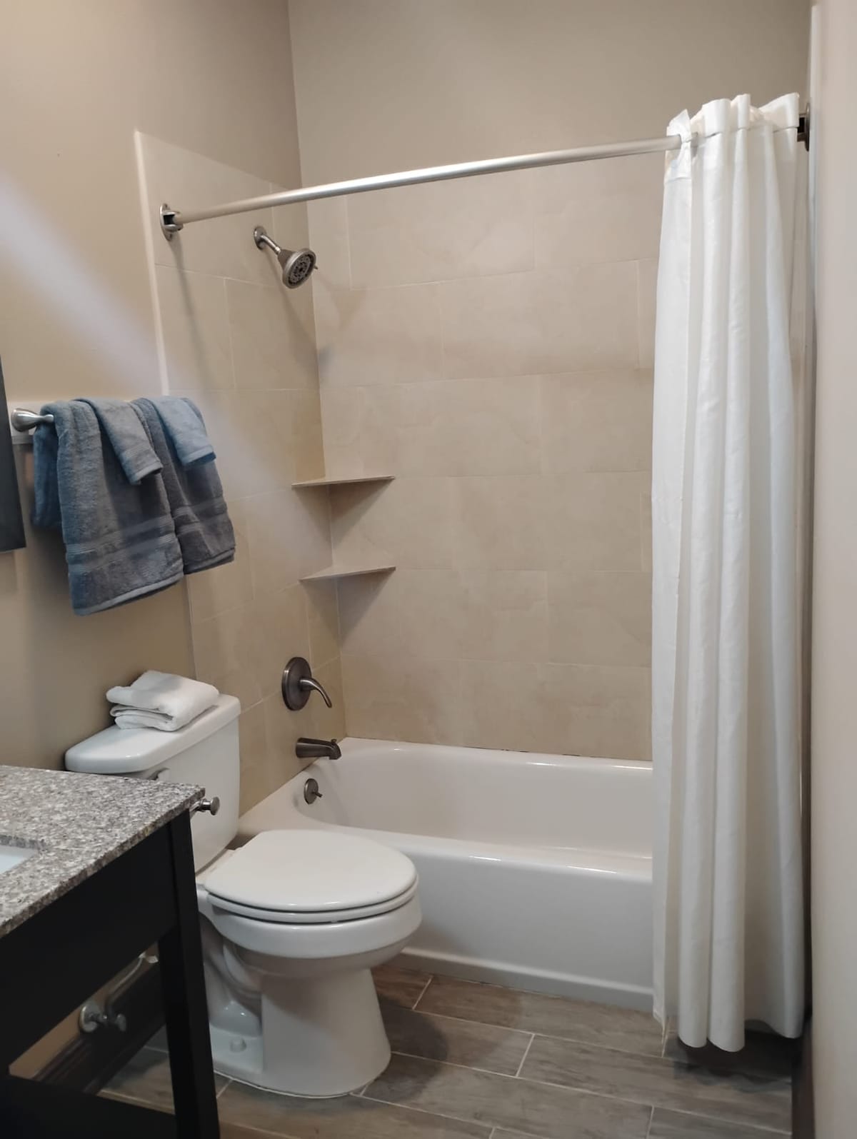 A neutral-toned bathroom features a bathtub with a shower curtain and a handheld showerhead. A sink with a granite countertop is positioned beside a toilet. Light-colored towels are neatly arranged on a rack, and additional toiletries are visible on a shelf.