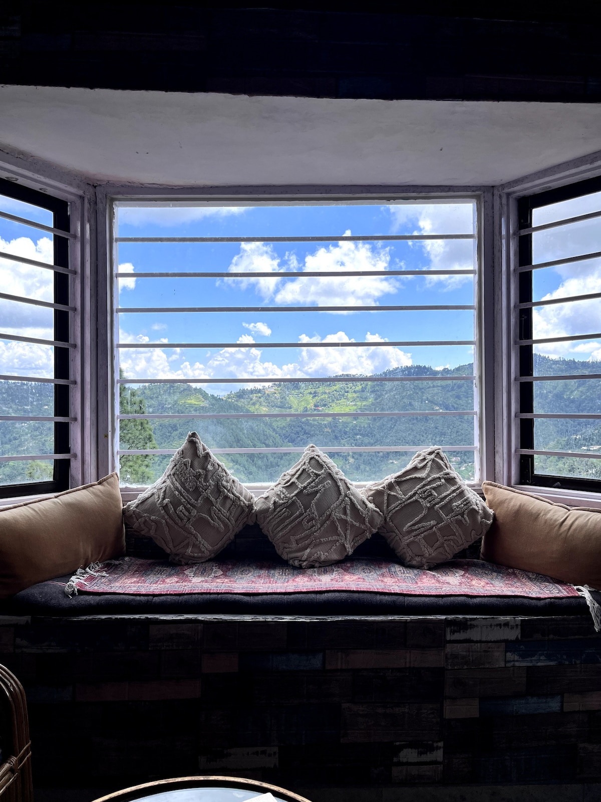 A comfortable window seat is positioned beneath expansive windows, offering views of the mountains and sky. The area features plush cushions and a decorative fabric draped across the seat. Natural light floods in, illuminating the inviting space.