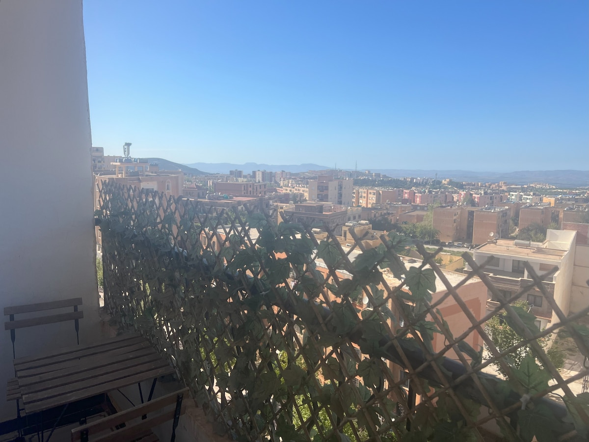 A panoramic view is presented from the balcony, featuring a lattice screen adorned with greenery. The landscape includes a spread of buildings and distant hills under a clear blue sky, offering a serene backdrop for relaxation.