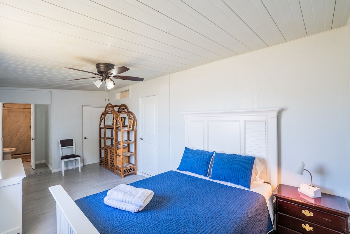 The spacious bedroom features a queen size bed with a blue quilt and white linens. A wicker chair and a stylish light fixture add to the decor. A ceiling fan is installed above. A walk-in closet and an adjoining bathroom are visible in the background.