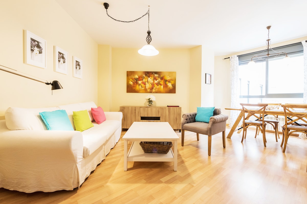 A contemporary living area features a white sofa adorned with colorful cushions. A wooden table stands beside a chair, and a wall-mounted lamp provides illumination. A decorative artwork is centered above a sideboard, with natural light filtering through large windows.