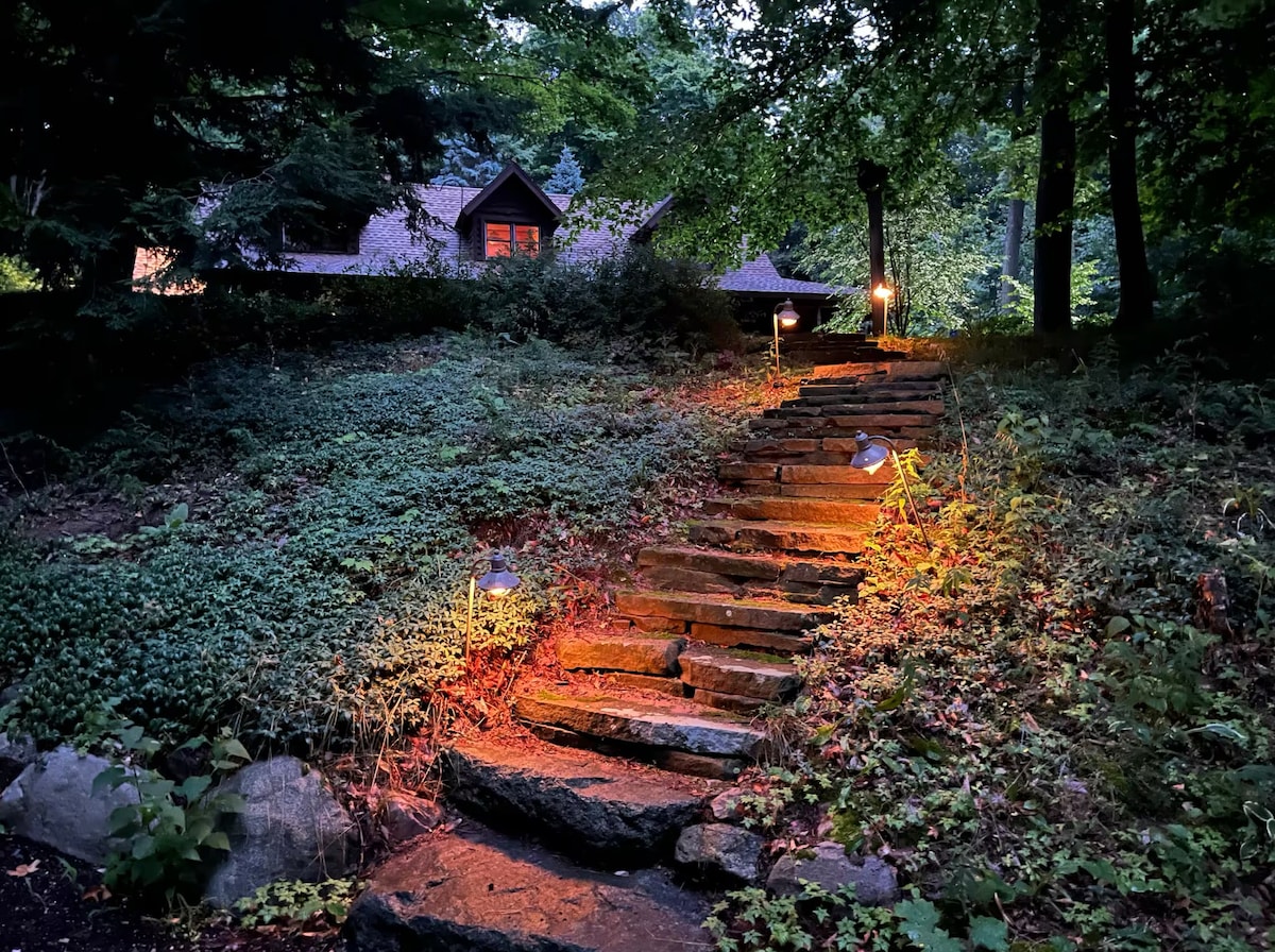 A stone pathway, illuminated by soft lighting, leads through lush greenery towards a cabin partially visible in the background. The surrounding trees create a natural setting, enhancing the inviting feel of the approach.