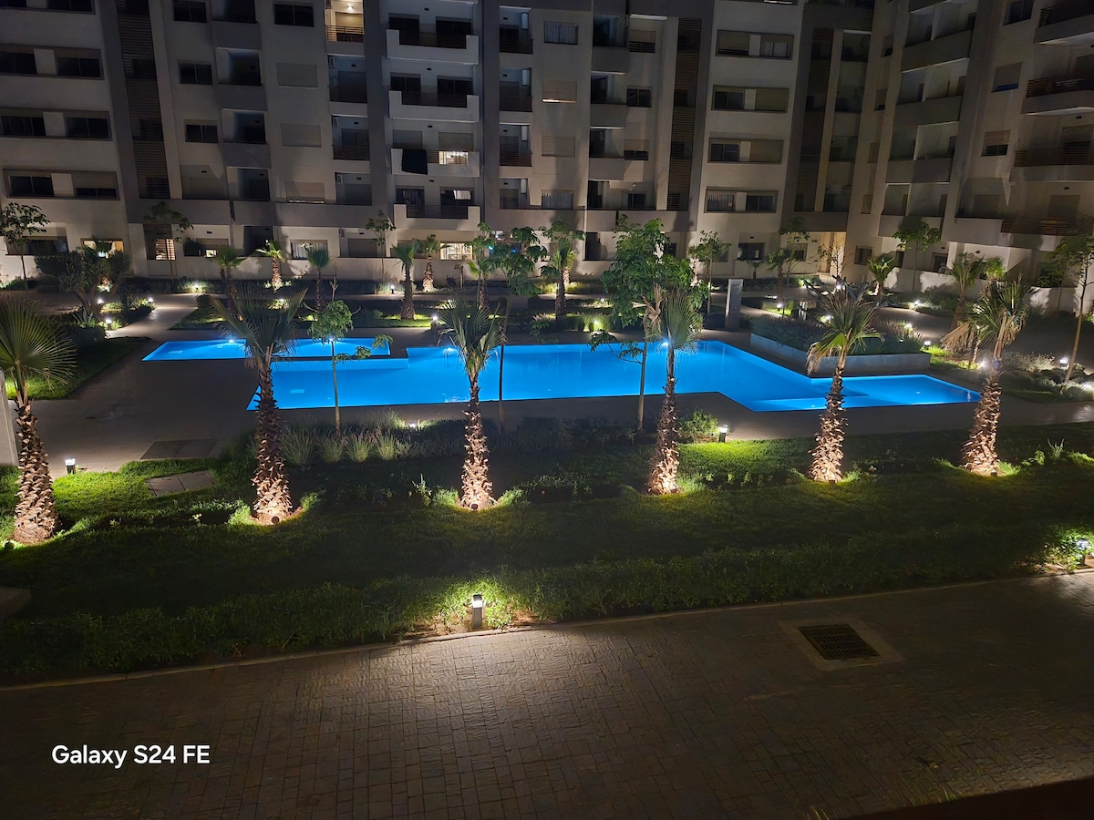 A spacious outdoor area is highlighted by a well-lit swimming pool, surrounded by palm trees and lush greenery. The pool's water glows in shades of blue, creating a serene ambiance. Modern buildings encase the area, adding a sense of privacy.