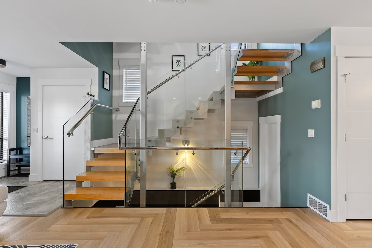 A modern staircase features a combination of hardwood steps and glass railings, illuminated by natural light from nearby windows. The walls are painted a calming blue, complemented by decorative artwork. Potted plants add a touch of greenery to the inviting entryway.