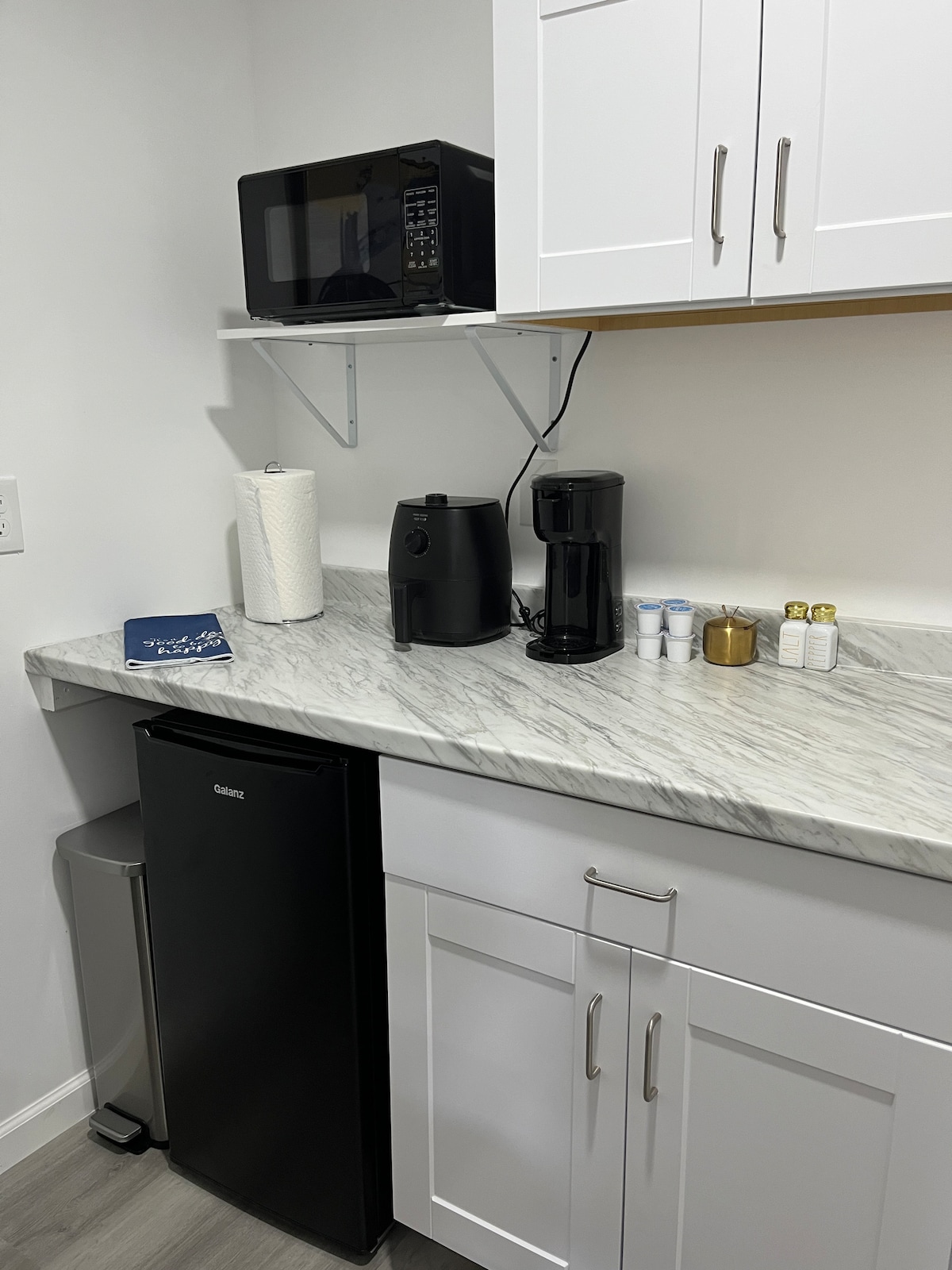 A kitchen space features modern white cabinetry and a marble countertop. A compact refrigerator is complemented by a microwave and coffee maker. Kitchen essentials such as paper towels and toiletries are neatly arranged, providing convenience for guests.