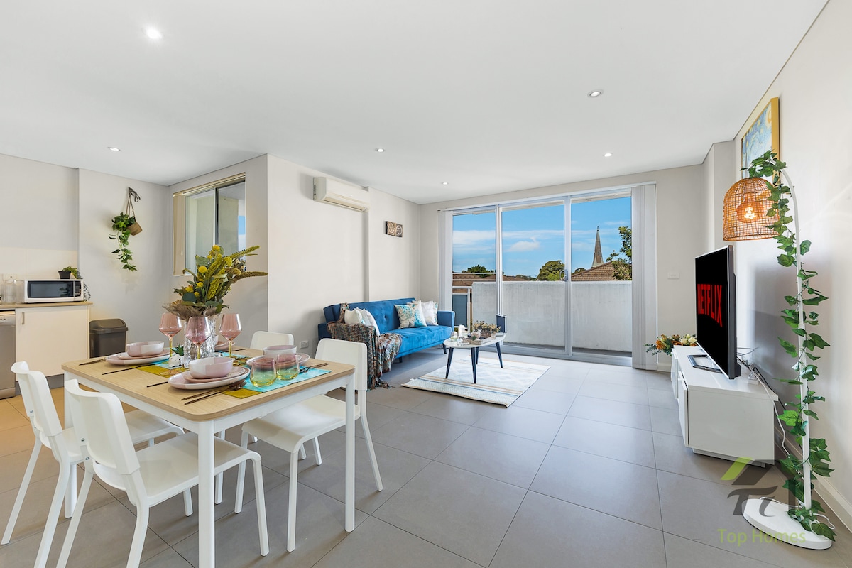 A modern living space is shown, featuring a dining area with a white table set for four and a comfortable blue sofa. Natural light fills the room through large windows that lead to a private balcony. A flat-screen TV and decorative plants add a contemporary touch.
