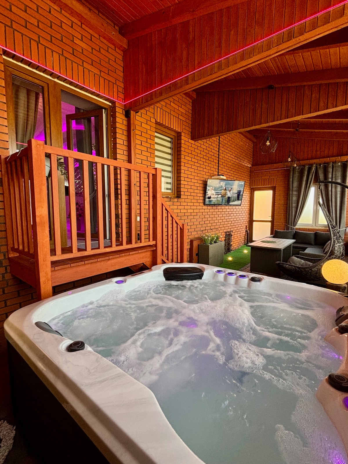 A private Jacuzzi SPA is visible with bubbling water. Nearby, a small balcony with wooden railing offers a view, and a wall-mounted TV is present. Comfortable seating is arranged, along with warm lighting accents and decorative elements within the brick-walled enclosure.