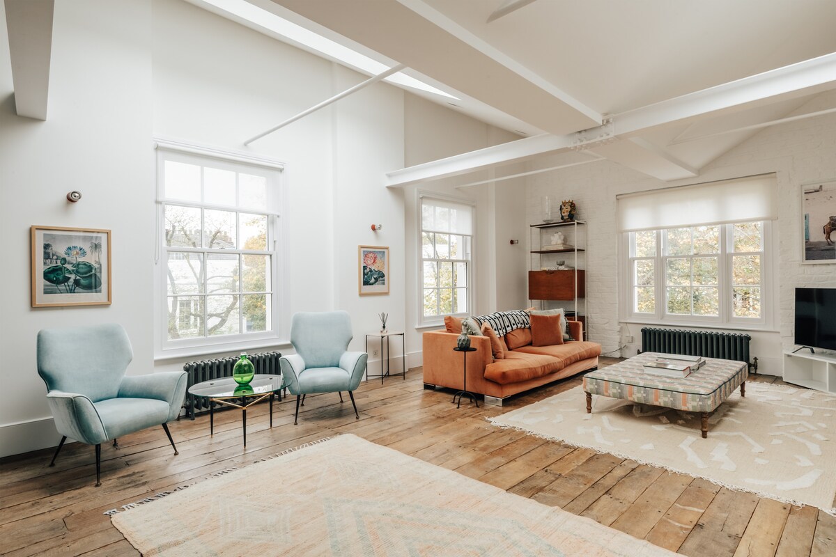 A spacious living area features large windows allowing ample natural light. Light-colored furniture includes a soft orange sofa and two blue armchairs, enhancing the room's design. A decorative coffee table sits in the center, accompanied by an area rug that adds texture to the reclaimed wood floors.