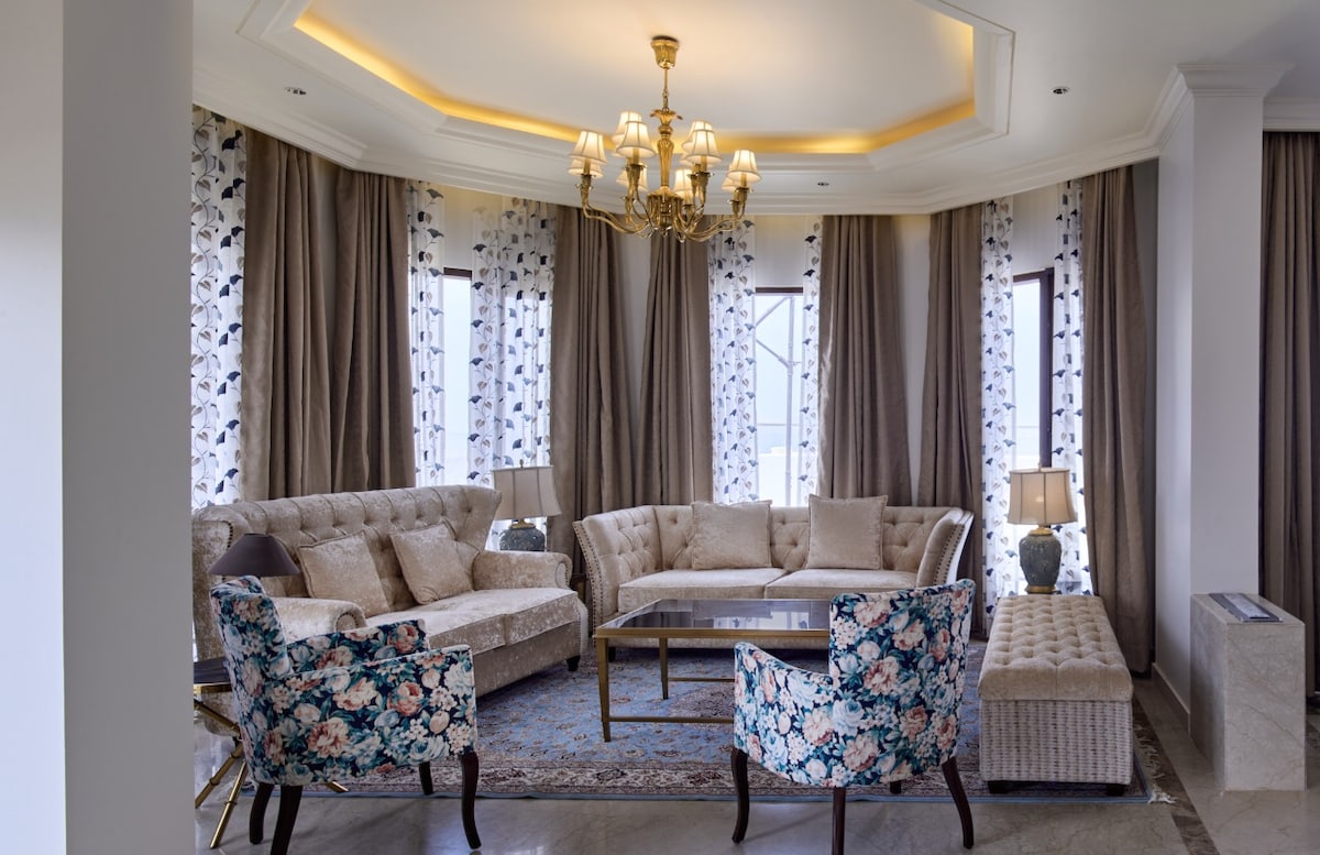 A spacious living area is presented with elegant furnishings, featuring a sectional sofa and a glass coffee table atop a decorative rug. Large windows are adorned with floral curtains, allowing natural light to fill the room, complemented by soft lighting from a chandelier above.