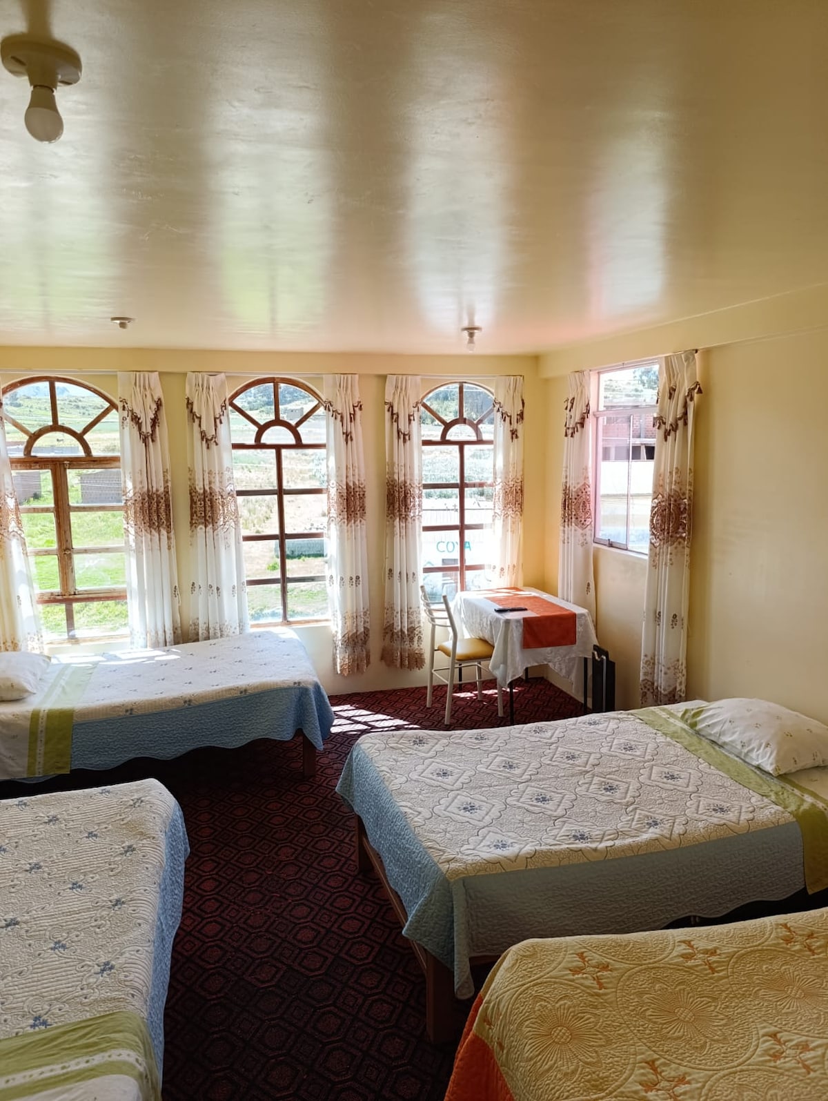 A well-lit room features four beds, each adorned with patterned bedding. Large windows are framed by light curtains, allowing natural light to fill the space. A small table and chair are placed near the window, offering a functional area for work or leisure.