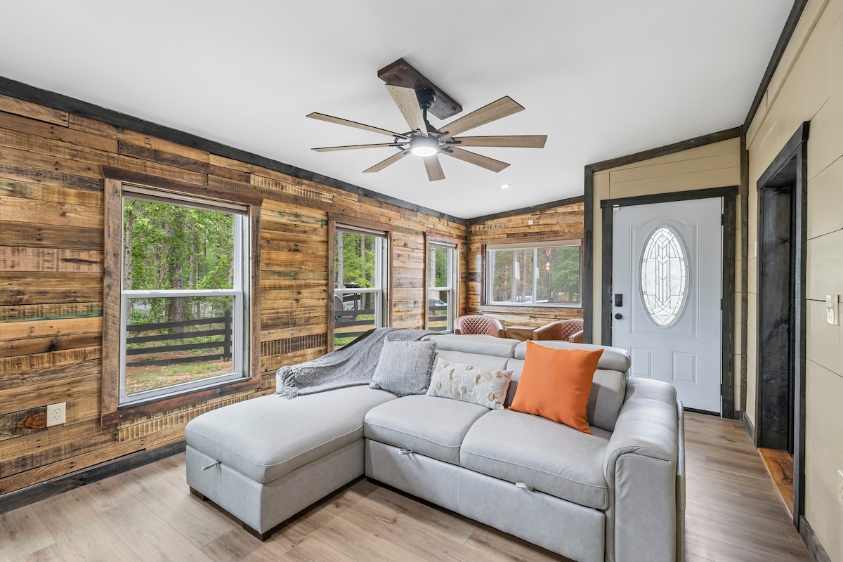 A welcoming living area features a large sectional sofa adorned with an orange accent pillow. Natural light enters through multiple windows, illuminating the rustic wood-paneled walls. A ceiling fan hangs overhead, contributing to the relaxing ambiance of the space.