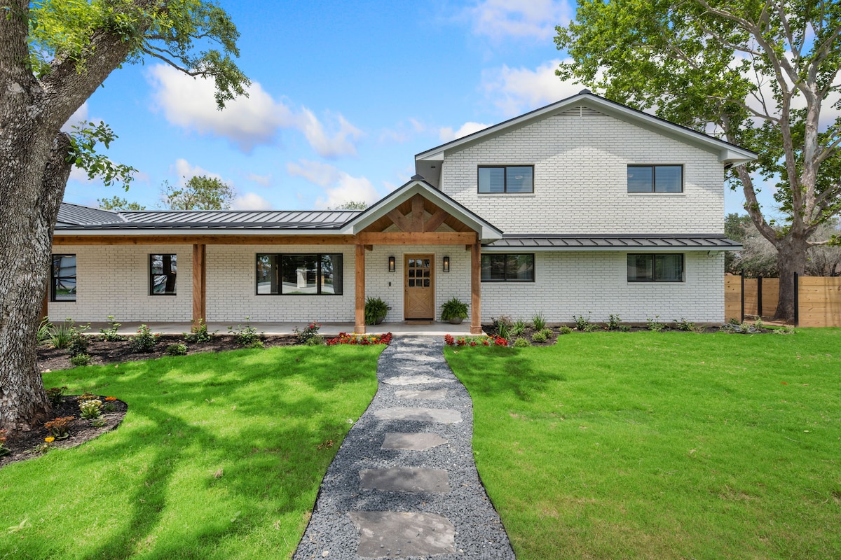 A modern farmhouse exterior is presented, featuring a well-maintained lawn and a stone pathway leading to the entrance. The structure is characterized by a combination of white brick and wood accents, surrounded by trees and neatly landscaped flower beds.