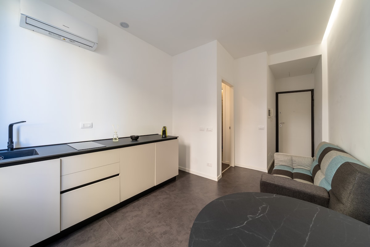 A modern kitchen area is presented with sleek cabinetry and dark countertops. An inviting sofa with a mix of grey and blue stripes is situated alongside a round table, creating a comfortable living space. Natural light is softly reflected on the smooth floor surface.