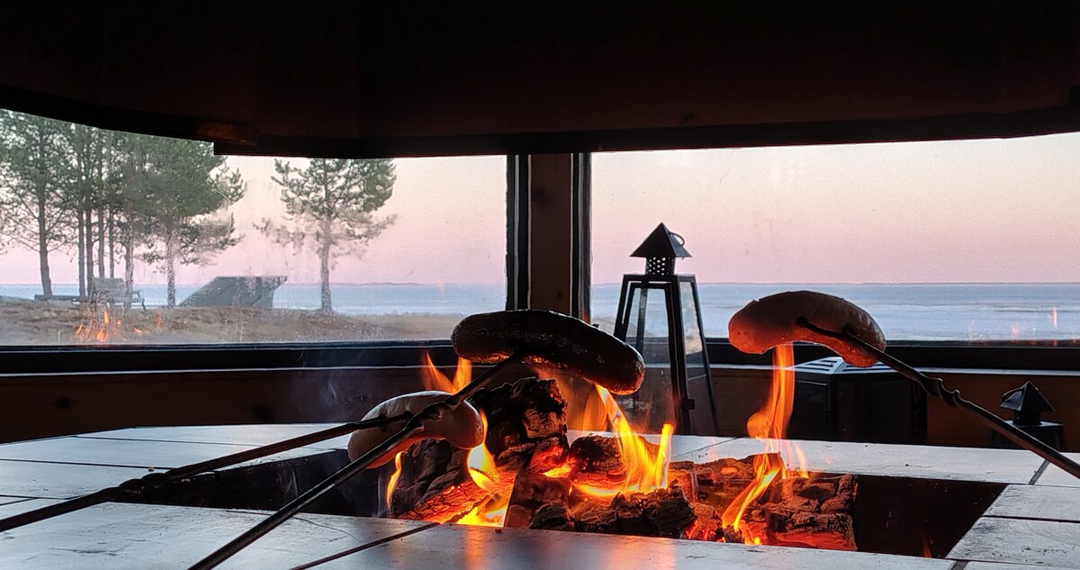 A cozy grill shelter features a central fire pit, with sausages skewered and cooking over the flames. Large windows provide views of the serene landscape, including trees and the water in the distance, with soft early morning light illuminating the scene.