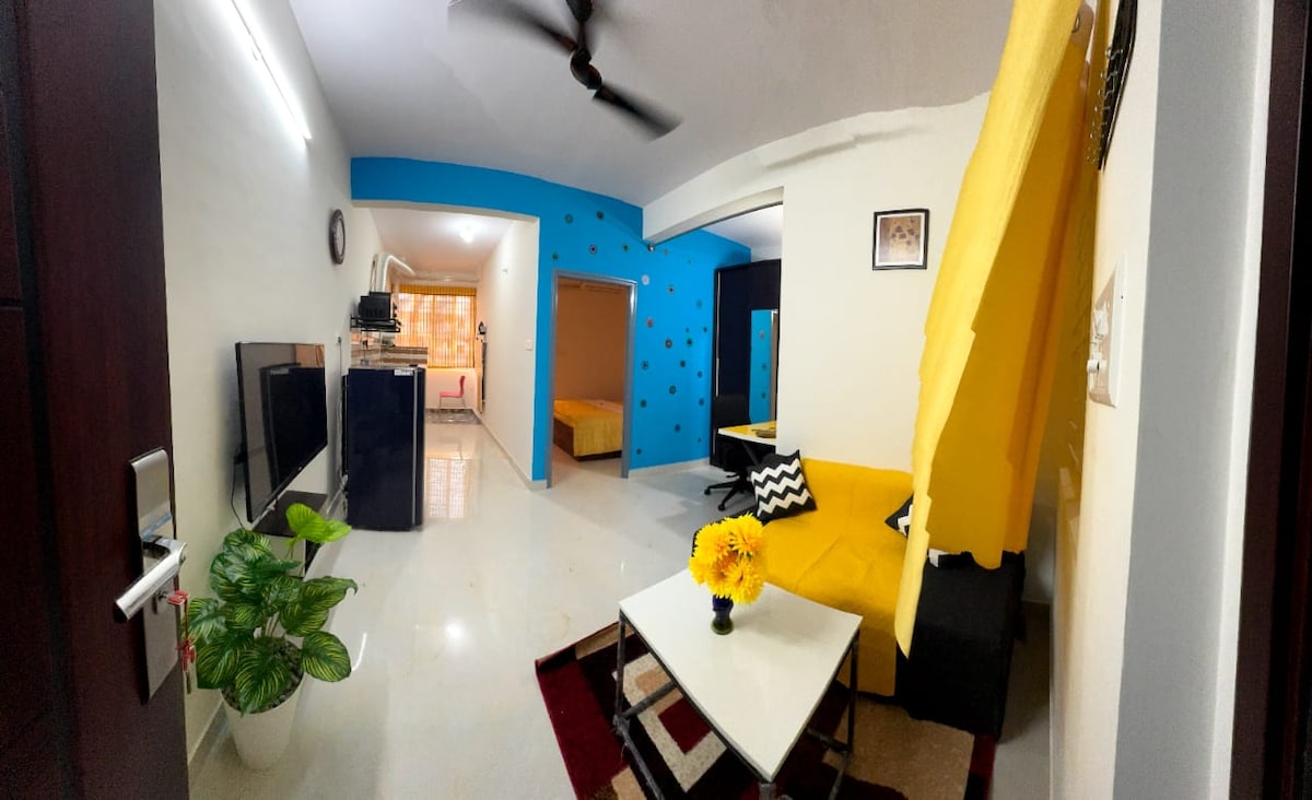 A vibrant living area features a bright yellow sofa paired with a minimalist coffee table. A decorative plant adds a touch of greenery. The walls are painted in calming shades, with a contrasting blue accent wall. Light from the adjacent kitchen contributes to the inviting atmosphere.