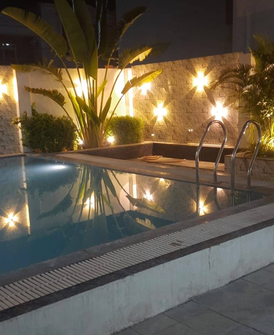 A private pool area is highlighted with decorative lighting that reflects softly on the water's surface. Lush greenery and modern stone walls frame the pool, while steps lead gracefully into the clear water, creating a serene ambiance for relaxation.