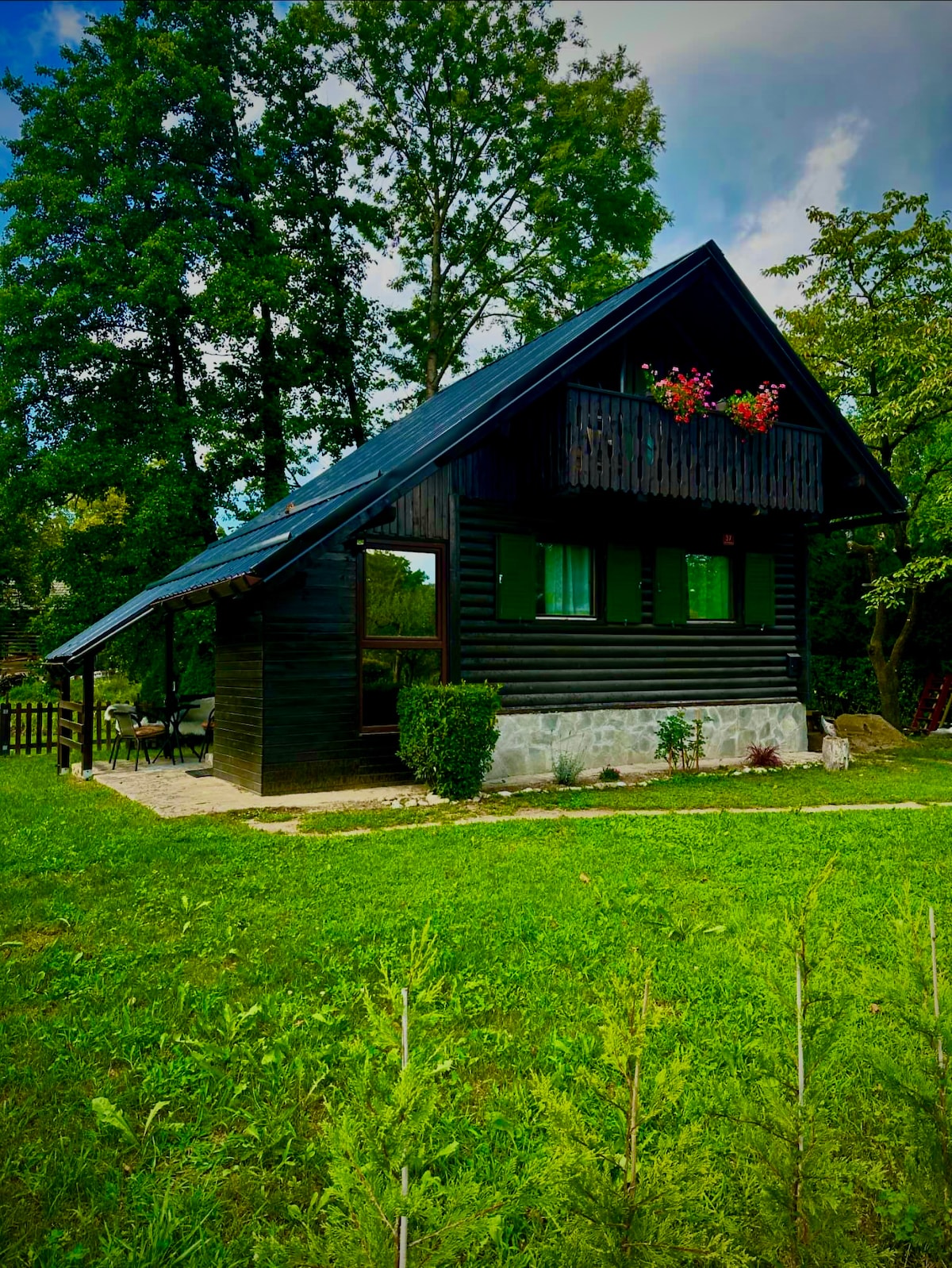 A charming cabin nestled in a lush green landscape features a steeply pitched roof and large windows. The front is adorned with colorful flowers in window boxes. A well-maintained pathway leads to a patio area, surrounded by trees that provide a natural backdrop.
