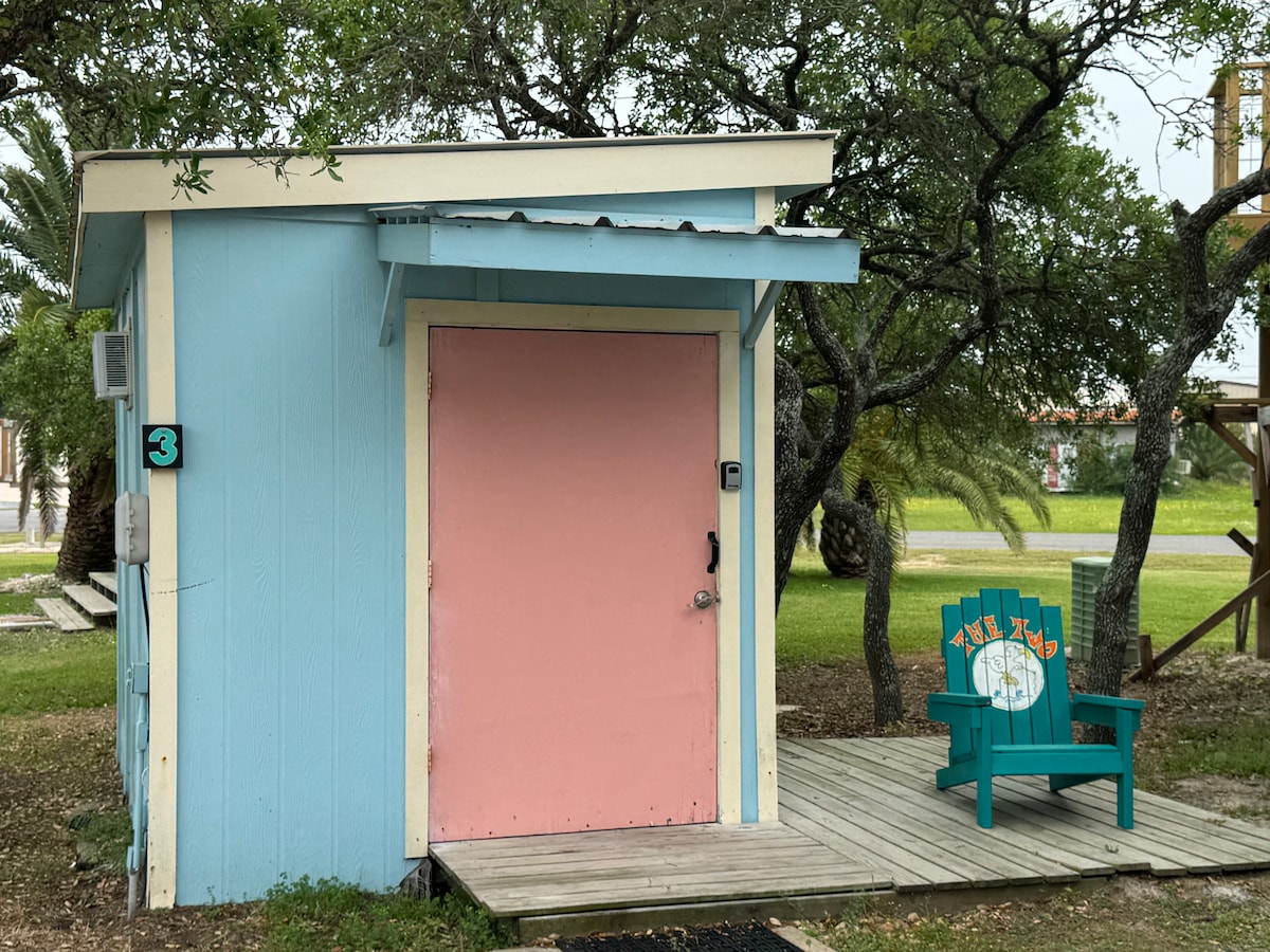 A colorful bunkhut features a light blue exterior with a contrasting pink door. A wooden deck leads to the entrance, complemented by a green chair displaying a friendly 'Chill Zone' sign. Surrounding greenery adds a natural touch to the inviting scene.