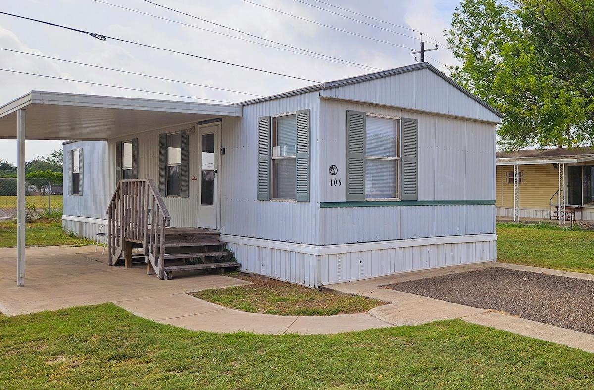 A single-story mobile home is presented with a light-colored exterior and a green accent line. The entrance features a set of wooden steps leading up to a front porch. A well-maintained grassy area surrounds the home, contributing to a serene outdoor setting.