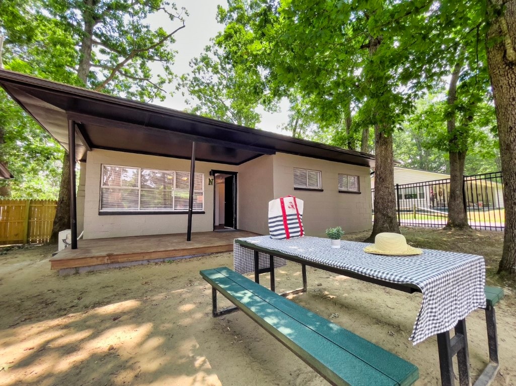 An outdoor seating area is featured, featuring a picnic table with a checkered tablecloth and a straw hat atop it. The cottage's exterior showcases large windows framed by trees, revealing a welcoming entrance.
