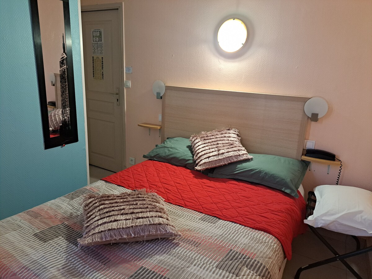 A cozy room features a spacious bed with red and green bedding, complemented by decorative pillows. A round wall light provides soft illumination. A mirror and a door can be seen on the light-colored walls, adding to the room's simplicity and functionality.