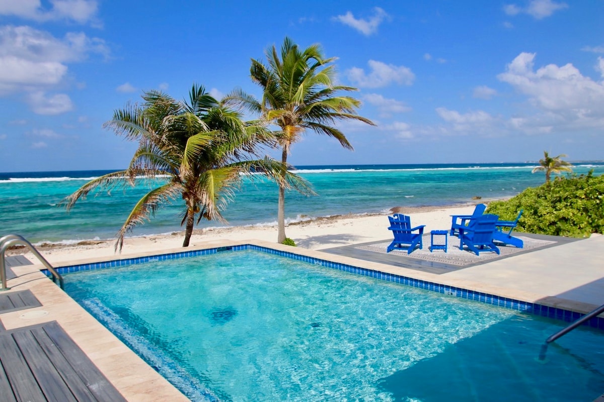 A private pool with a crystal-clear surface reflects the bright blue sky. Palm trees provide shade nearby, while vibrant blue outdoor chairs are positioned on a deck overlooking the soft sandy beach and the turquoise waters of the Caribbean Ocean.