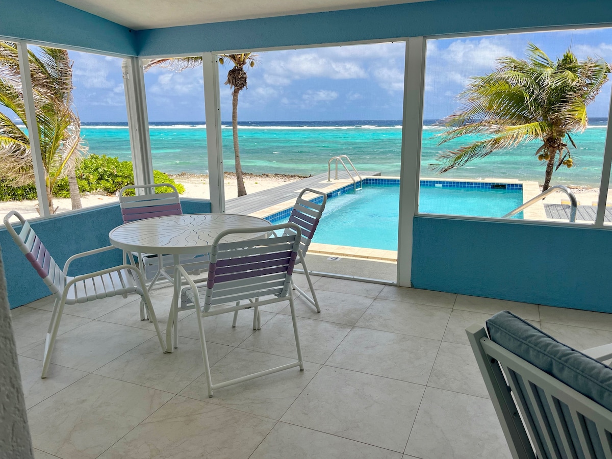 A spacious screened porch is furnished with a round dining table and several chairs, offering views of the turquoise waters. Sunlight filters through the glass, highlighting the private pool and nearby sandy beach framed by palm trees.