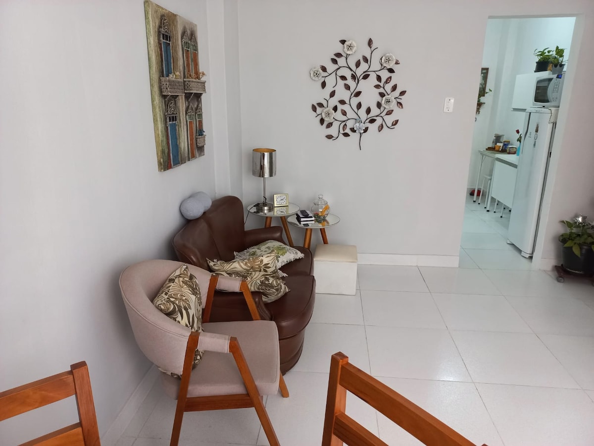 A cozy sitting area features a brown armchair and a light-colored chair, adorned with decorative cushions. A small table is positioned beside the brown chair, and a wall art piece depicting a tree branch is mounted nearby. A glimpse of the equipped kitchen can be seen in the background.