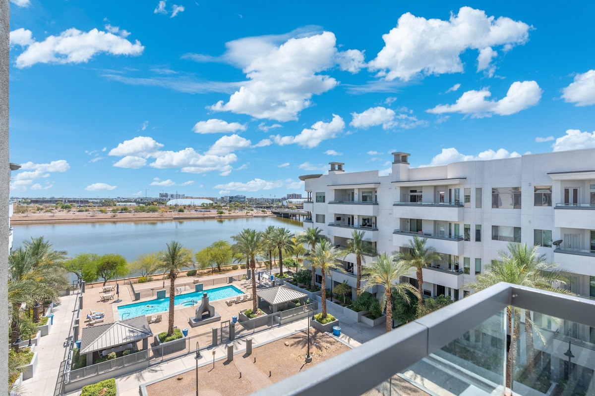 An expansive view capturing a lake and a community pool area below, surrounded by palm trees. The modern building features clean lines and balconies, reflecting a bright blue sky dotted with clouds.