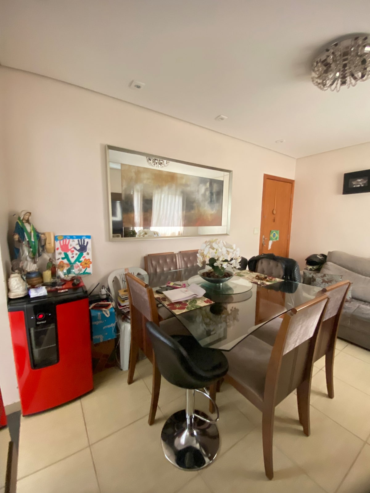 A dining area features a glass table surrounded by several brown chairs. Decorative items and a white floral arrangement are present on the table. A red water cooler stands nearby, and a mirror reflects the space, adding depth. A subtle chandelier hangs from the ceiling.