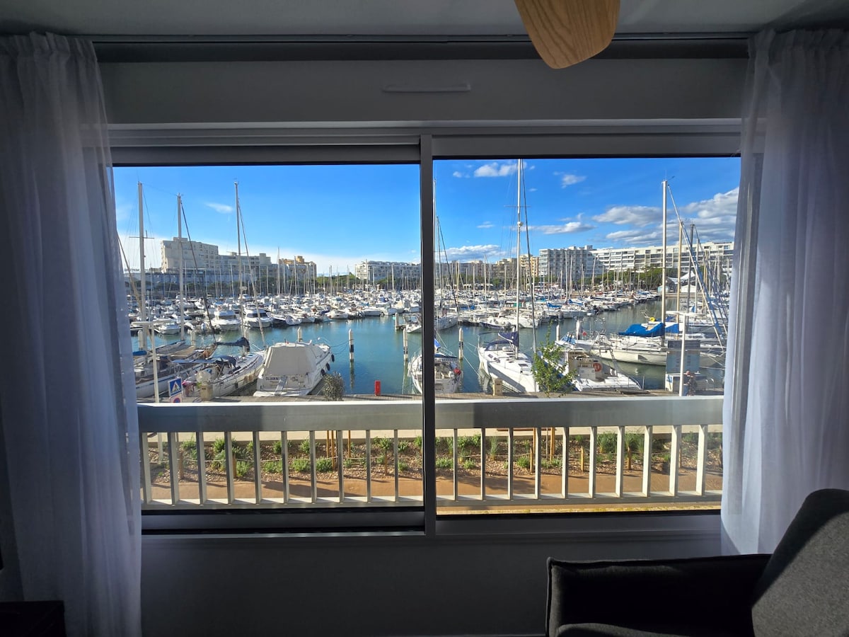 A panoramic view of the marina is captured through a large window, showcasing numerous boats docked in the water. Bright blue skies and scattered clouds add to the serene atmosphere, while sheer curtains frame the window, allowing natural light to enhance the interior space.