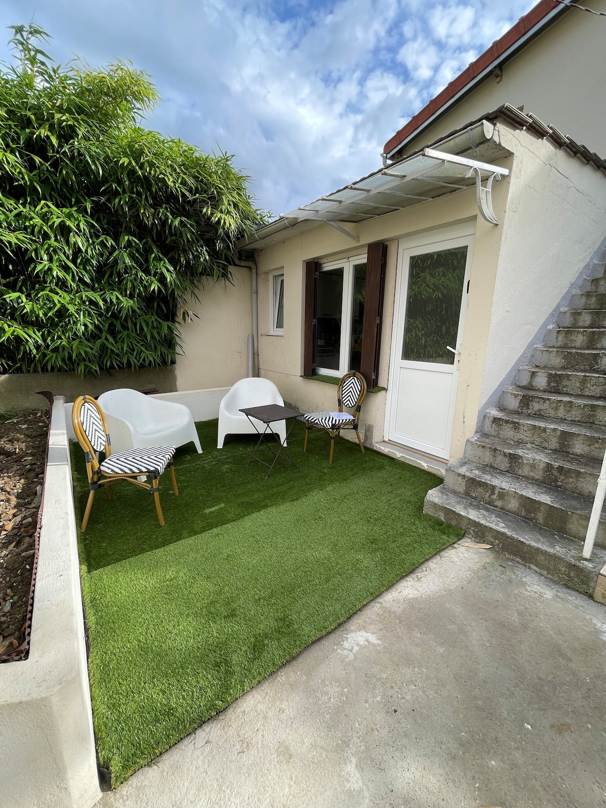 A small outdoor space is featured with a grassy surface, including two patterned chairs and a small table. A staircase leads to the entrance of the adjoining building, while lush greenery is visible in the background under a partly cloudy sky.