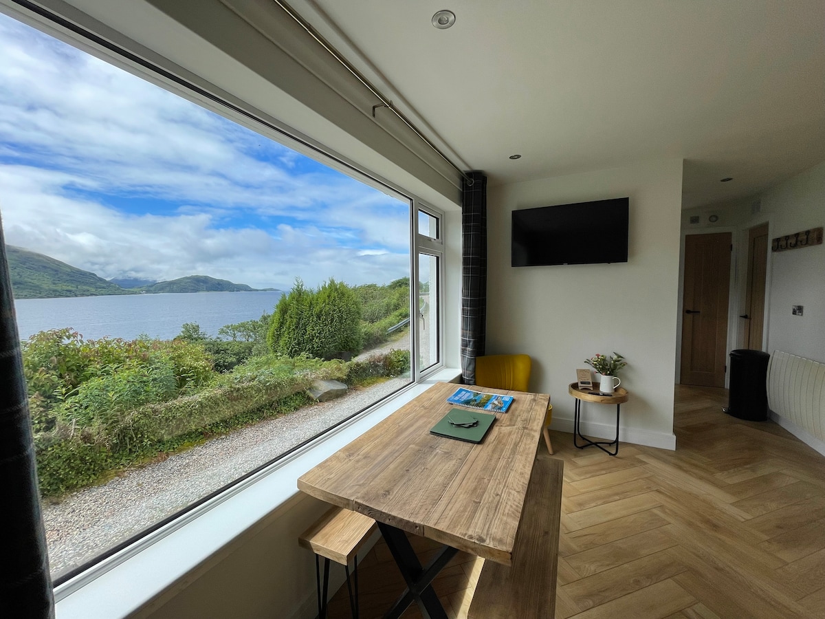 A dining area features a wooden table with matching benches, positioned beside a large window that showcases views of Loch Linnhe and the surrounding hills. A small television is mounted on the wall opposite, and a yellow accent chair adds a pop of color.