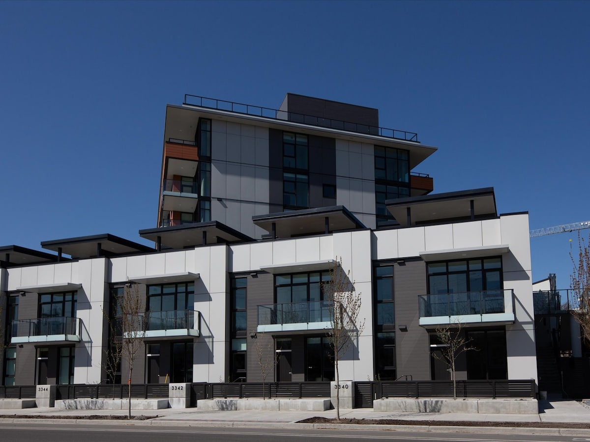 A modern townhome complex features multiple units with large glass doors and balconies, allowing natural light to enter. The building has a clean, contemporary design with a combination of gray and white panels. A clear blue sky serves as the backdrop.