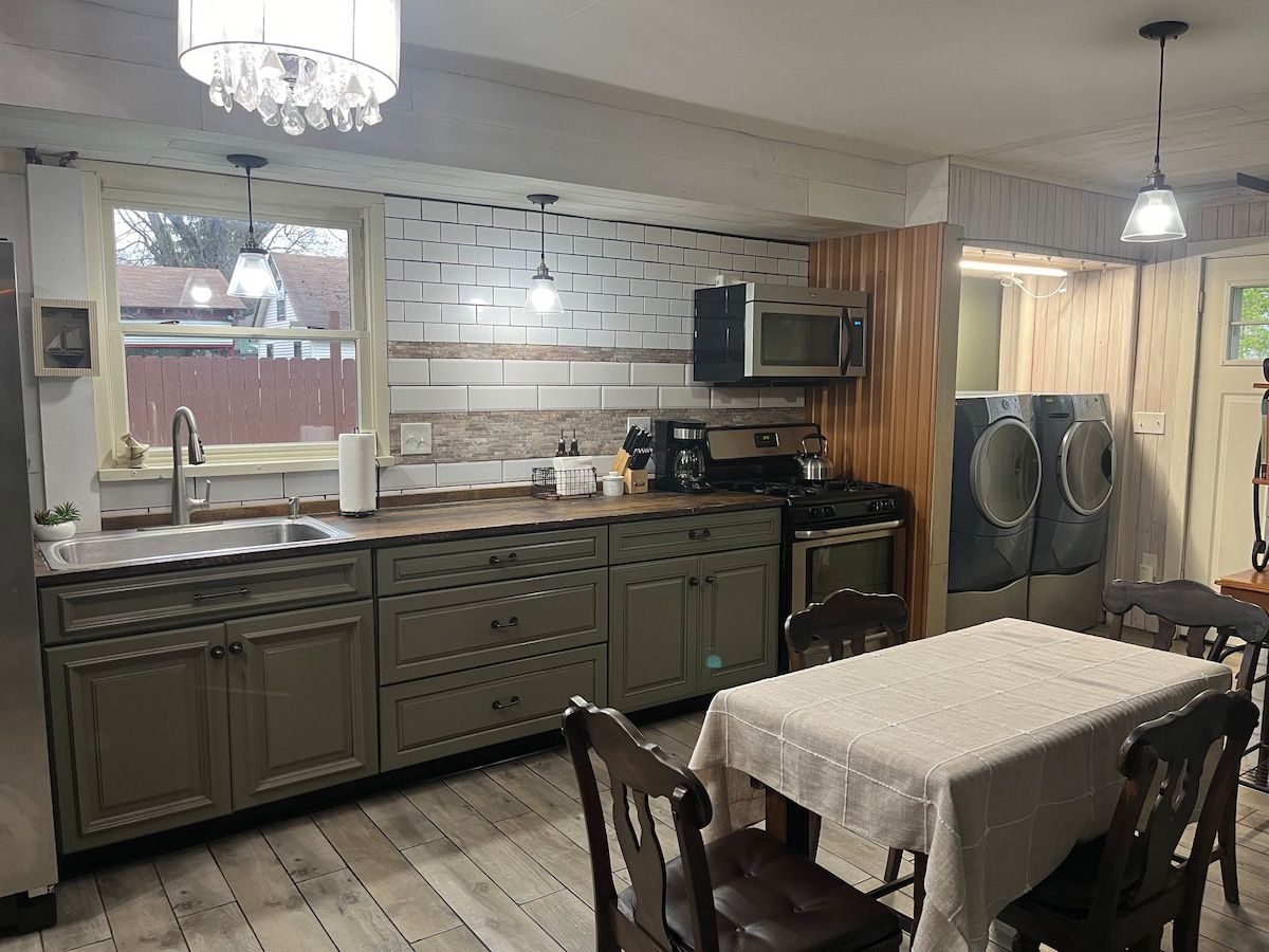 The kitchen features modern appliances, including a microwave and gas stove, set against a charming backsplash. Natural light streams through the window above the sink. A dining table sits nearby, and a washer/dryer is conveniently located in the adjacent space.