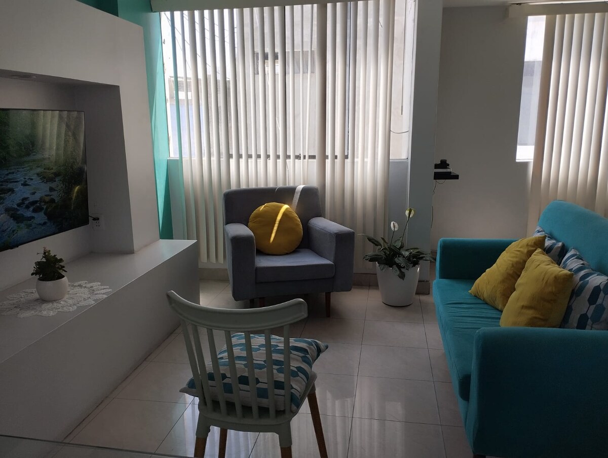 A cozy living area features a blue sofa and a light blue armchair with a decorative yellow pillow. A television is mounted on the wall, and natural light filters through the vertical blinds. A potted plant adds a touch of greenery to the space.
