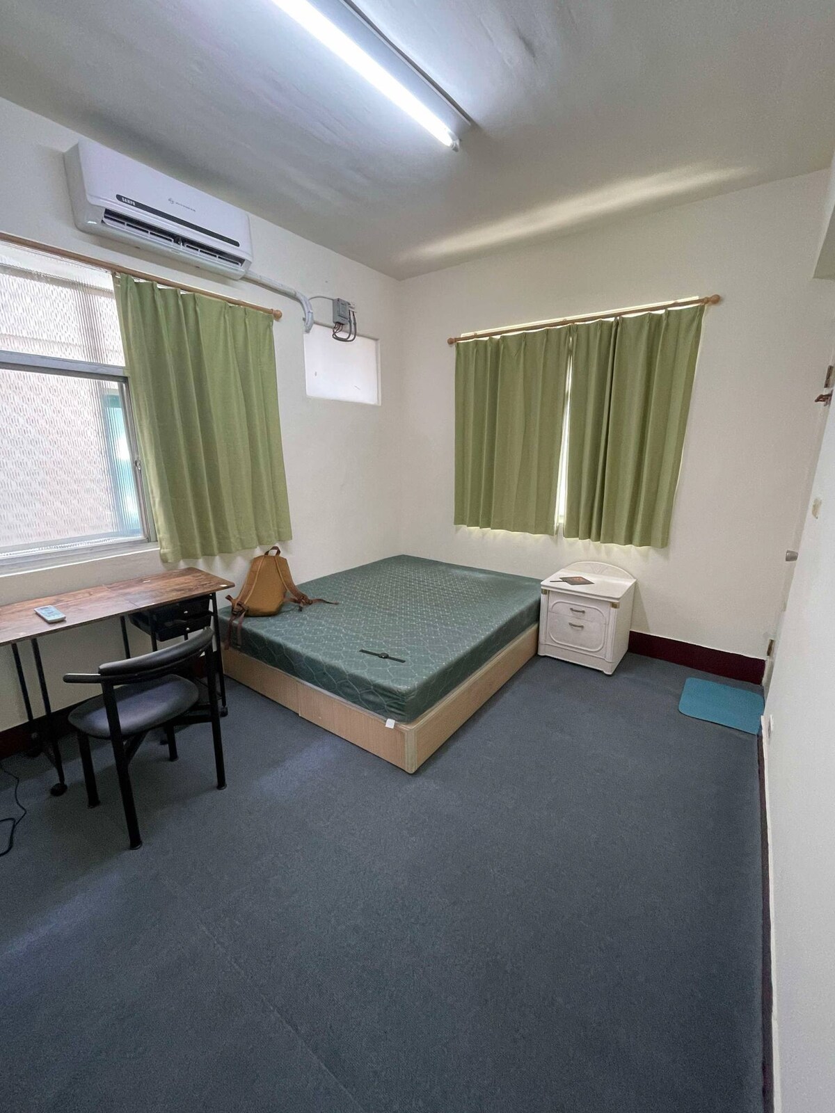 A simple bedroom is furnished with a spacious bed, a small desk with a chair, and a nightstand. Two green curtains frame the window, allowing natural light to fill the room. A compact air conditioning unit is mounted on the wall above.