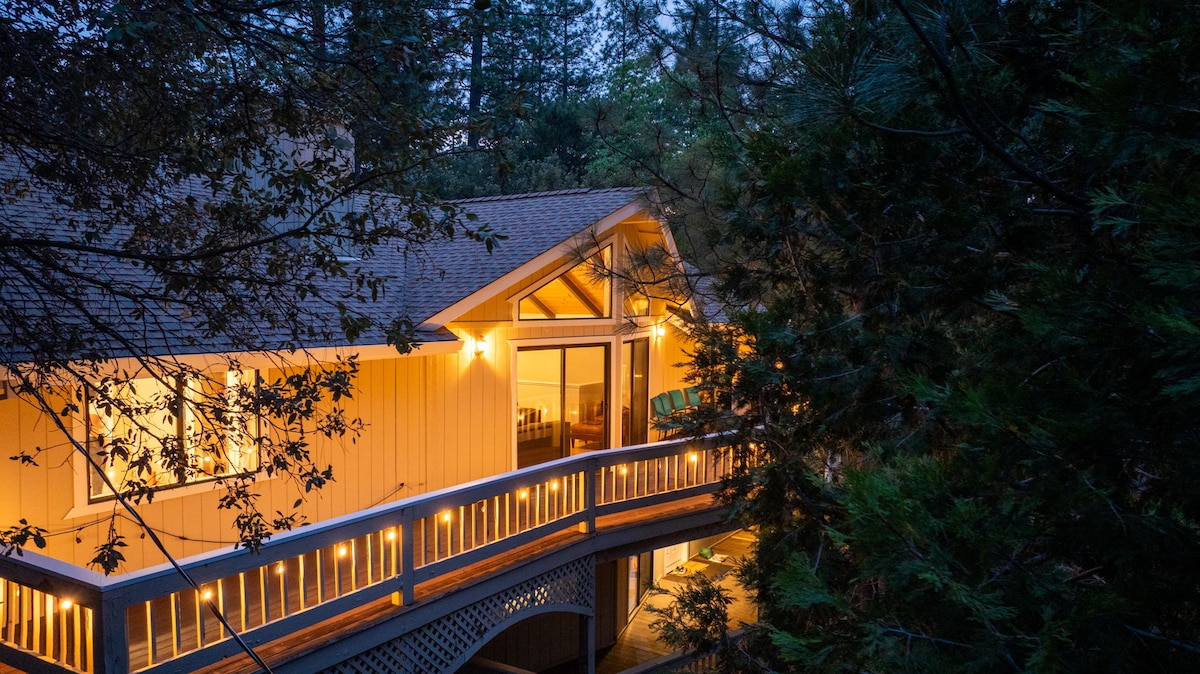 The exterior of the Whispering Pines Lodge is framed by surrounding trees, with warm lights illuminating the home from the expansive deck. Large glass doors invite views of the interior, enhancing the welcoming atmosphere as twilight settles in.
