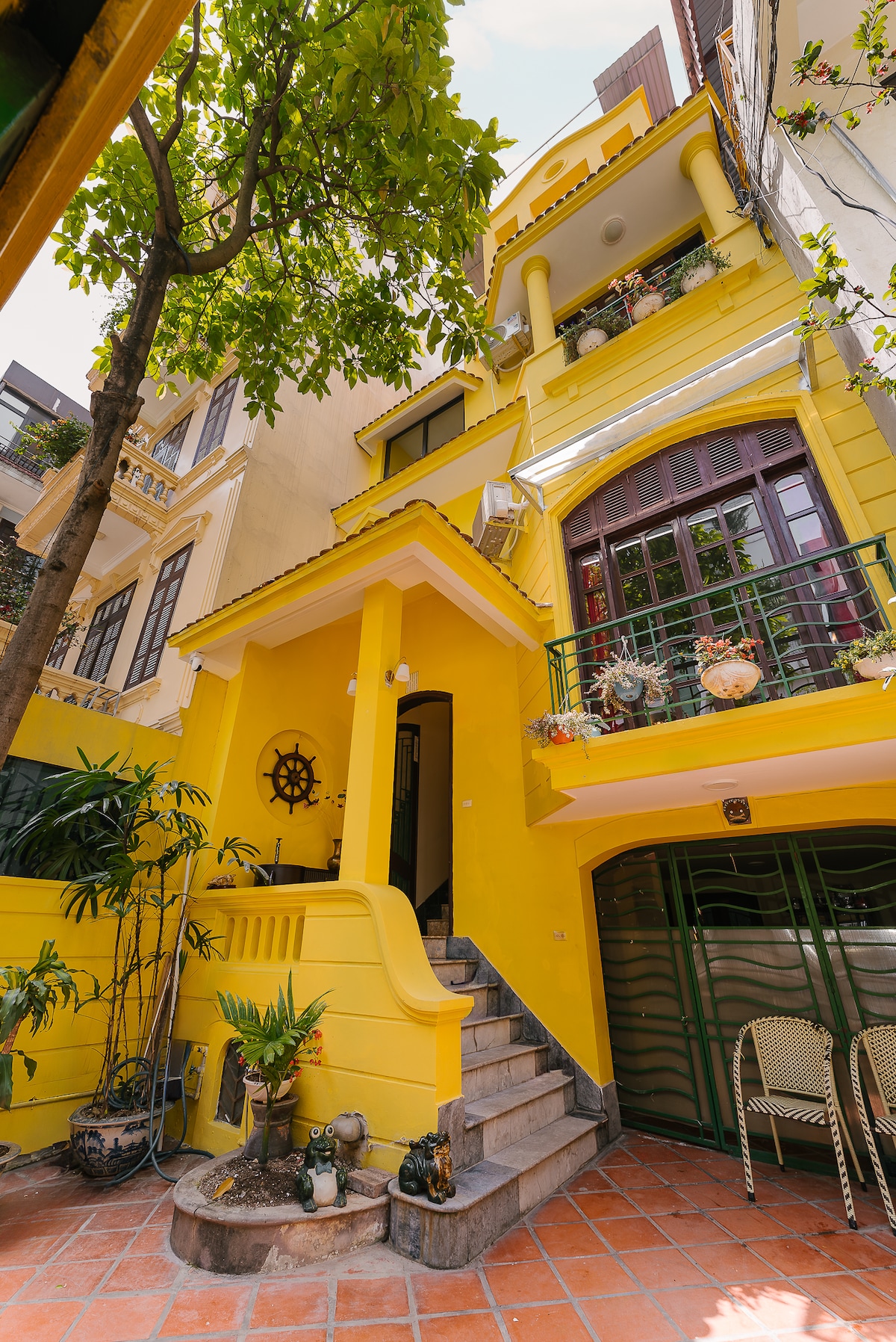 A vibrant yellow exterior of the villa is showcased, surrounded by greenery. The entrance features sculpted stairs leading to an inviting doorway. Decorative elements, such as plants and balconies, add character, while terracotta tiles cover the ground, creating a warm atmosphere in the courtyard.