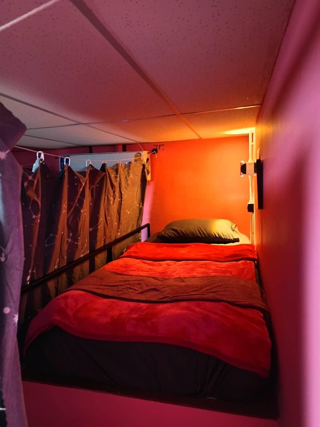 A snug sleeping nook is framed by soft fabric curtains, providing a sense of privacy. The space features a compact bed dressed in warm red and dark bedding, with gentle lighting highlighting the vibrant pink walls.