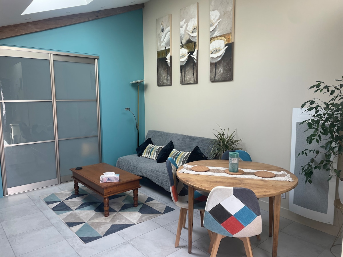 A living area features a comfortable grey sofa adorned with decorative cushions, paired with a wooden coffee table. A round dining table with colorful chairs is visible, alongside a geometric patterned rug. Two large floral artworks are displayed on the wall, enhancing the environment.
