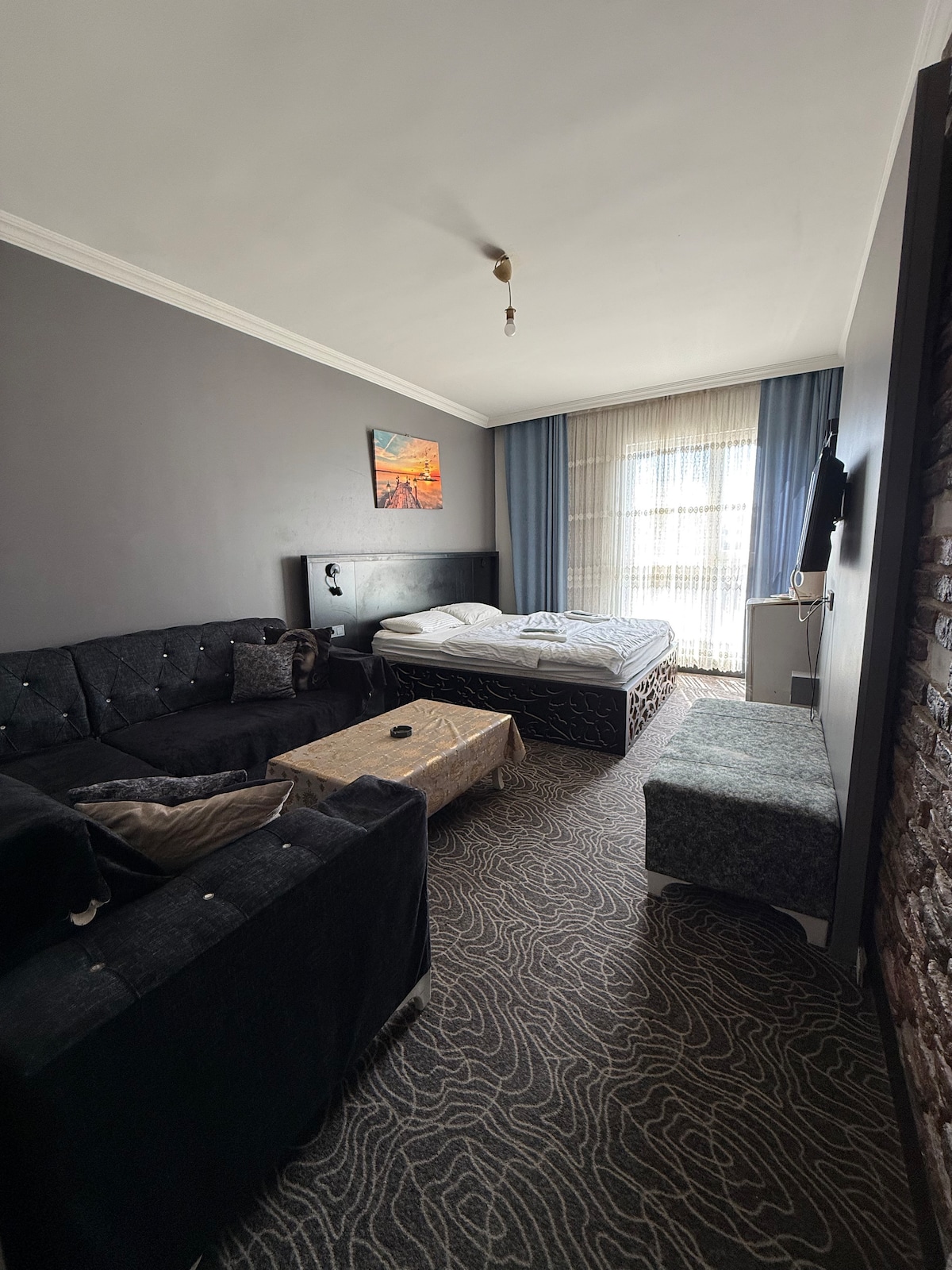 A comfortable room features a spacious bed with white bedding and a modern couch. A coffee table is positioned in front of the couch, while a textured ottoman adds additional seating. Natural light filters in through the large window adorned with sheer curtains.