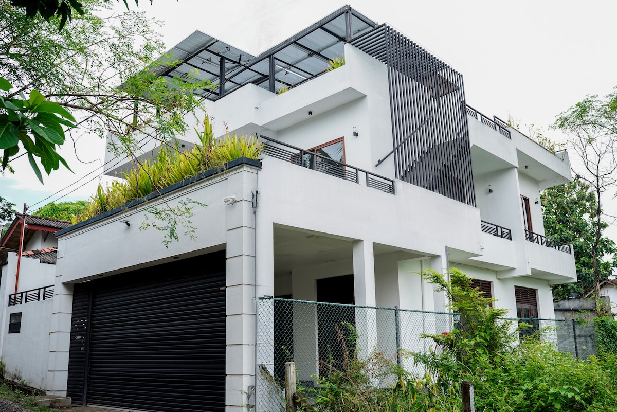 The modern building features a sleek white façade and multiple levels, enhanced by lush greenery on the roof. A protective fence surrounds the property, and a garage door is visible at the front. Large windows allow for natural light, creating a bright and open feel.