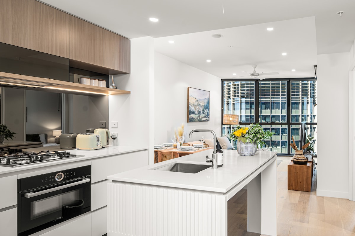 The modern kitchen features sleek cabinetry and a spacious island with a breakfast bar. Natural light floods the area through large windows, showcasing views of the vibrant living space. A gas stove, microwave, and colorful decor elements enhance the inviting atmosphere.