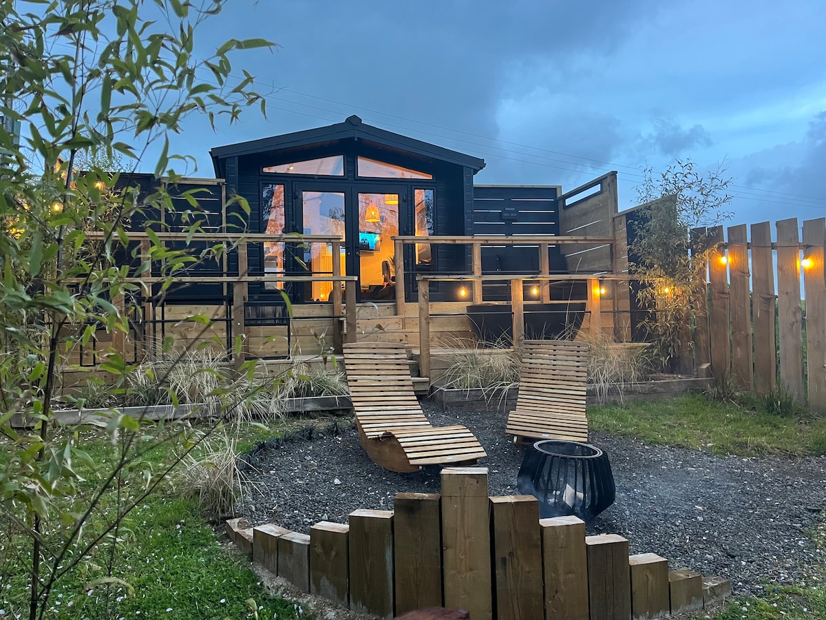 A rustic cabin is visible behind a welcoming outdoor space, illuminated by soft lights. Curved loungers are arranged around a circular fire pit, surrounded by natural landscaping. The cabin features large windows, allowing light to pour in, and is bordered by wooden fencing.