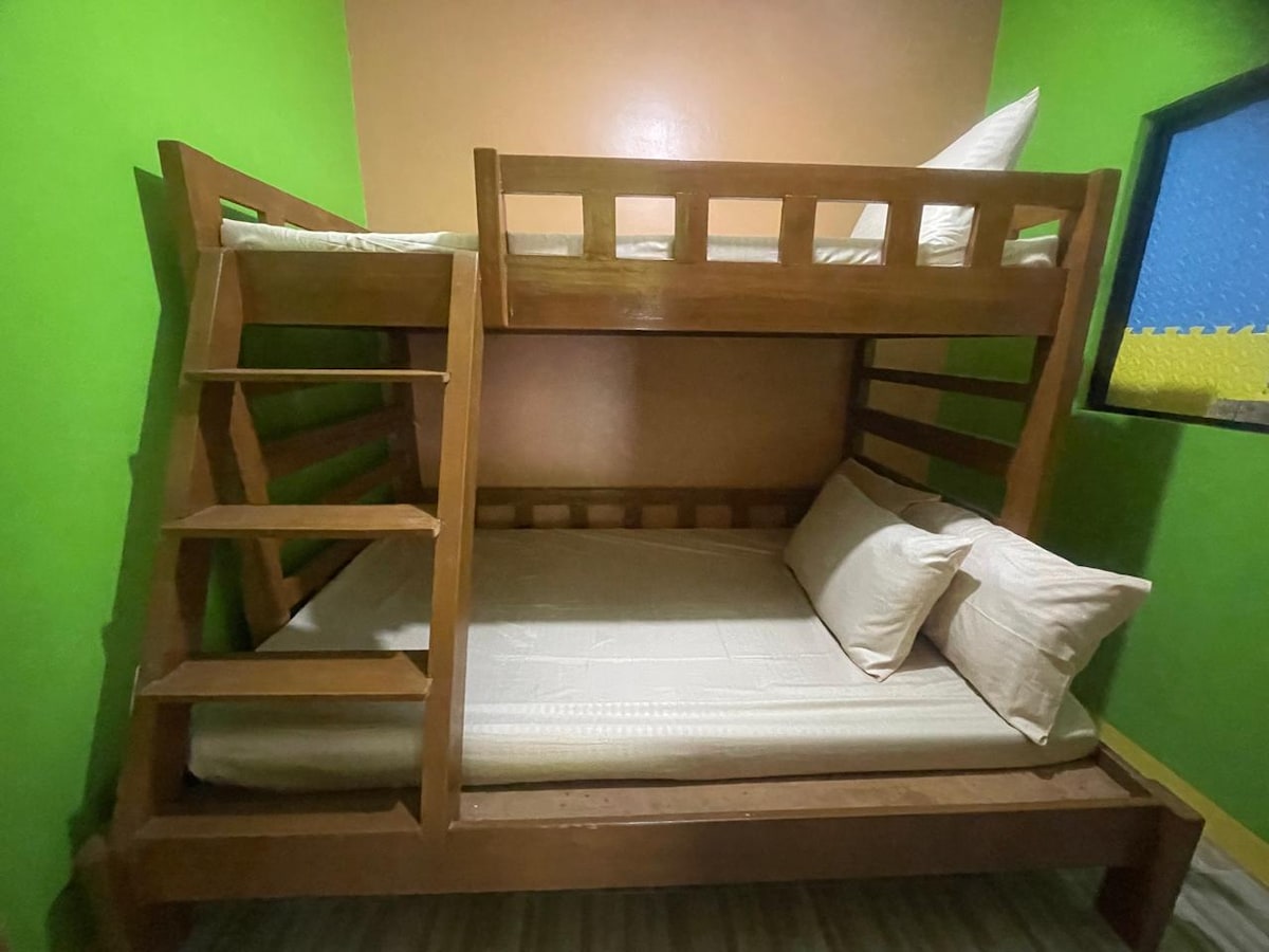 A wooden bunk bed is featured in a cozy sleeping area, with a double bed on the bottom and a twin bed on top. Soft, neutral-colored bedding complements the vibrant green walls. Two pillows rest on the bottom bed, providing comfort and inviting rest.