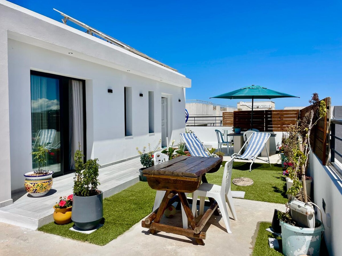 A private rooftop space is presented, featuring a wooden dining table surrounded by potted plants and flowers. Two striped loungers are positioned for relaxation. An umbrella provides shade over a seating area, while green grass adds a touch of nature, enhancing an inviting outdoor atmosphere.