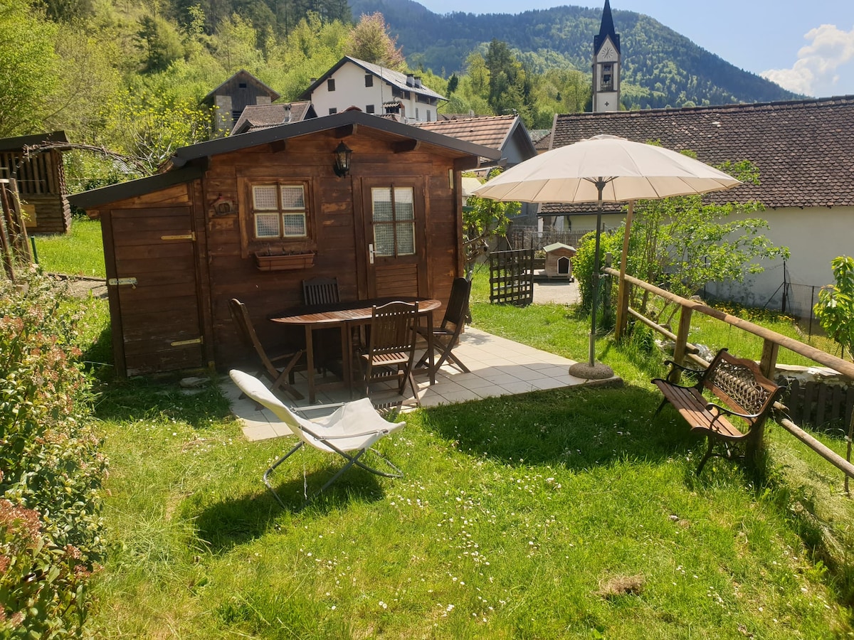 A wooden garden cabin is surrounded by lush greenery, featuring a patio table with four chairs and a large umbrella for shade. A pair of outdoor lounge chairs and a decorative bench are positioned nearby, all situated on a grassy area with scenic mountain views in the background.