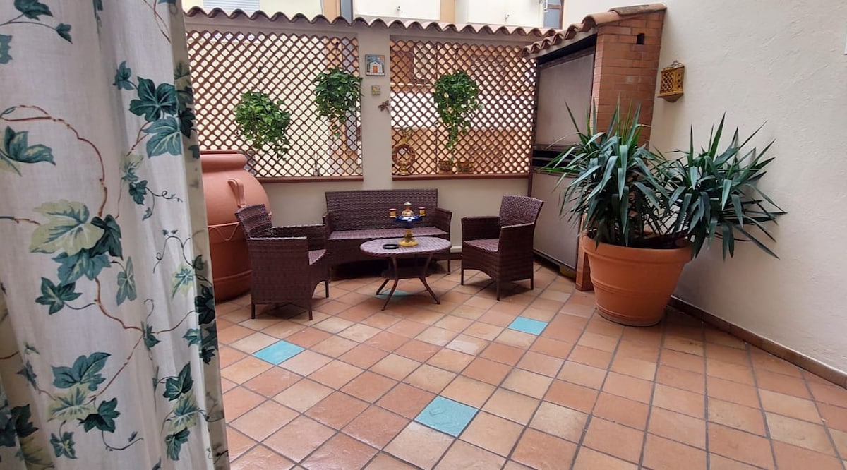 An outdoor courtyard is presented with terracotta flooring and decorated with greenery. A small round table is positioned at the center, surrounded by two wicker chairs. A larger potted plant adds a natural element to the space, while a patterned curtain drapes softly in the background.