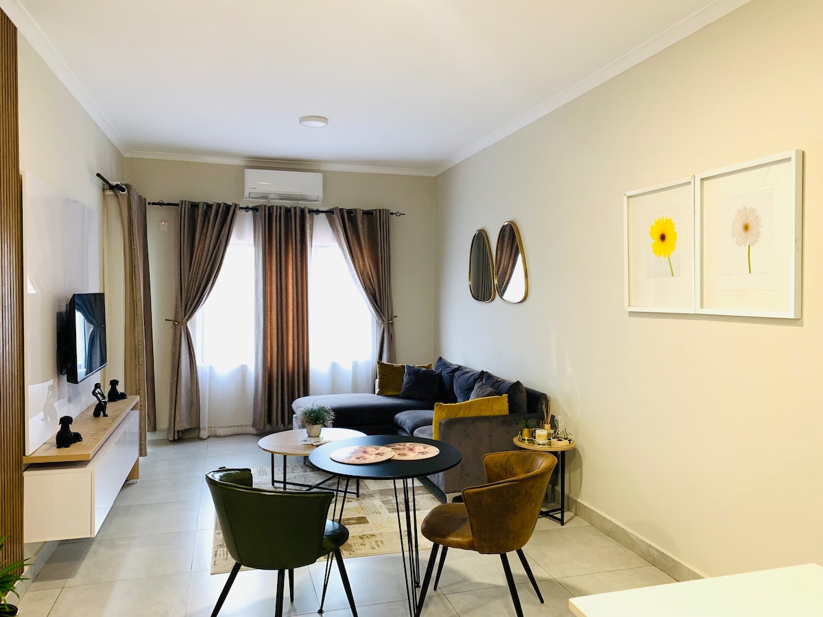 The living room features a comfortable sofa, complemented by two upholstered chairs. A round table is positioned in the center, with decorative items adorning the surface. Natural light filters through two large windows, dressed in soft curtains, enhancing the modern color palette of the space.